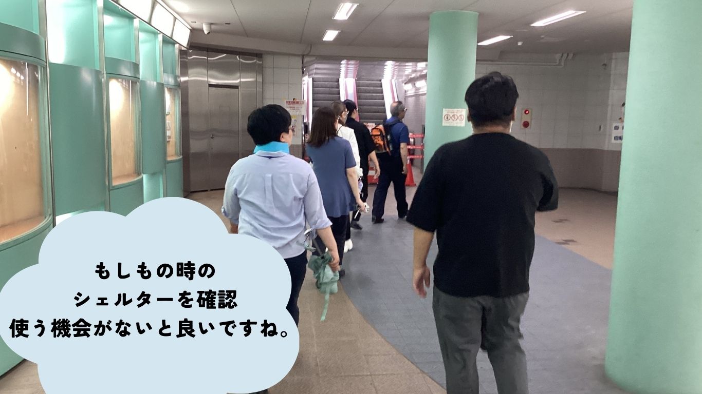 Subway station hallway with several people walking; a large pale-blue speech bubble in the lower-left shows Japanese text about checking emergency shelters.