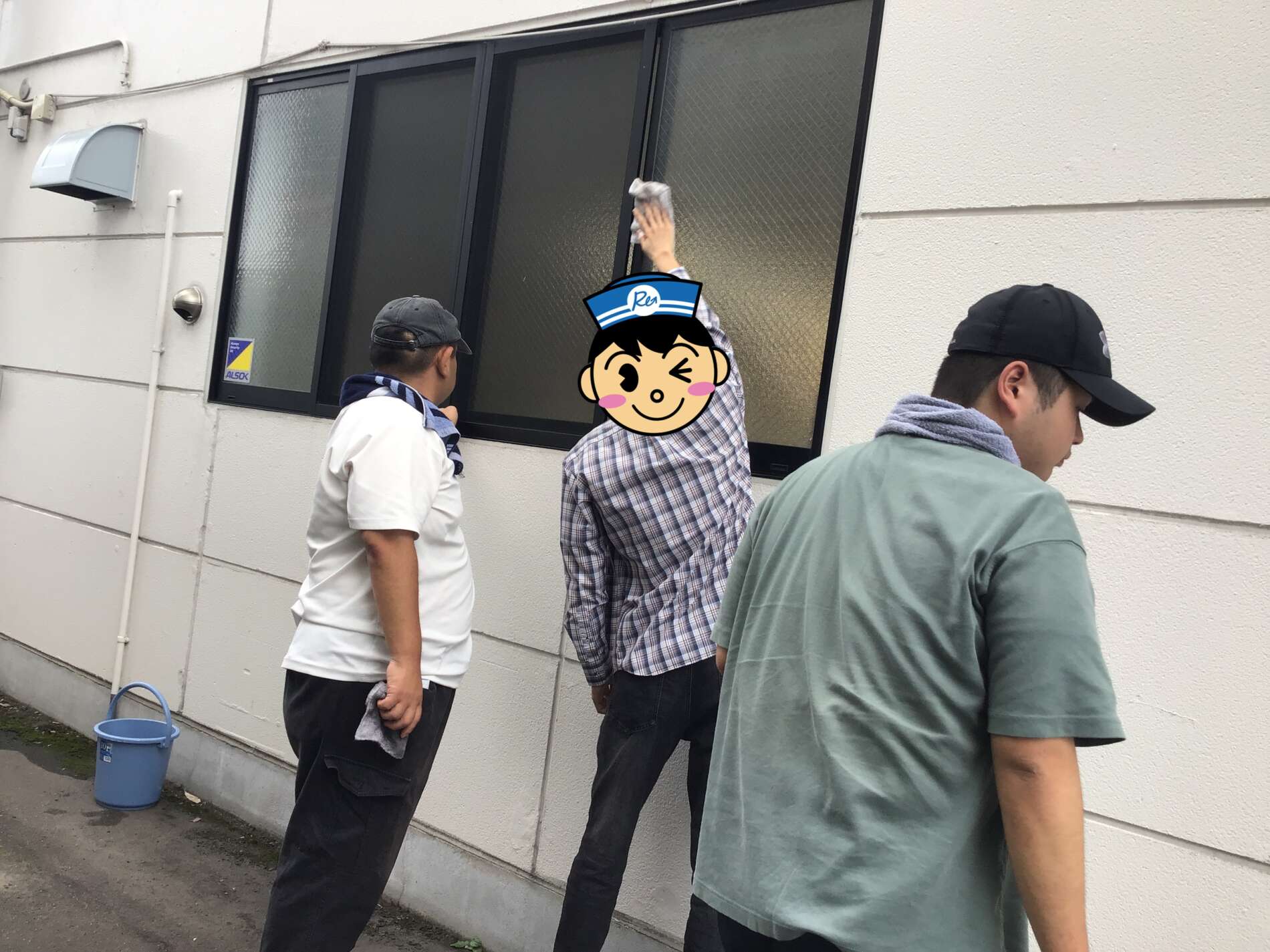 Three men stand outside a beige building by a large frosted window; one raises a gloved hand, with a cartoon sticker covering his face.