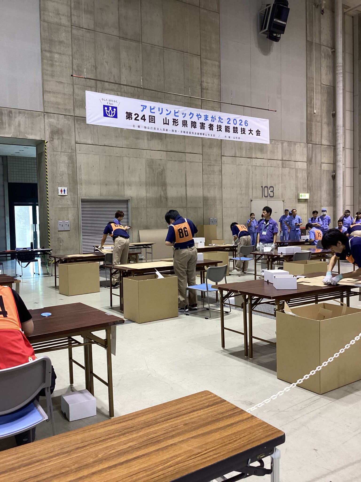 Indoor hall with people in numbered vests assembling tables and cardboard boxes for an event; banner in Japanese overhead reads a 2026 competition