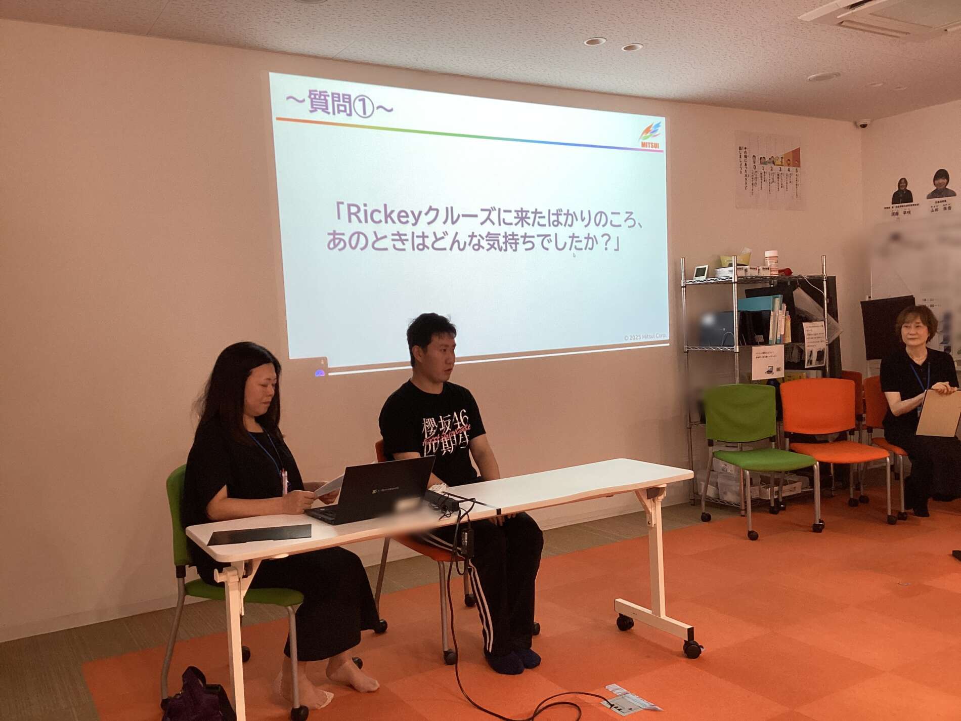 Panel discussion in a classroom: three people seated at a white table with laptops, a projected slide with Japanese text on the wall behind them.