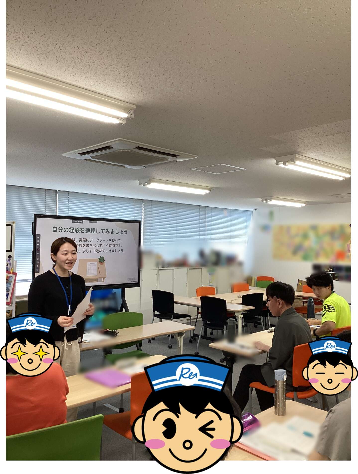 Instructor standing at front of classroom presenting to adults around tables; large display screen shows Japanese text. Cartoon avatars partially cover foreground.