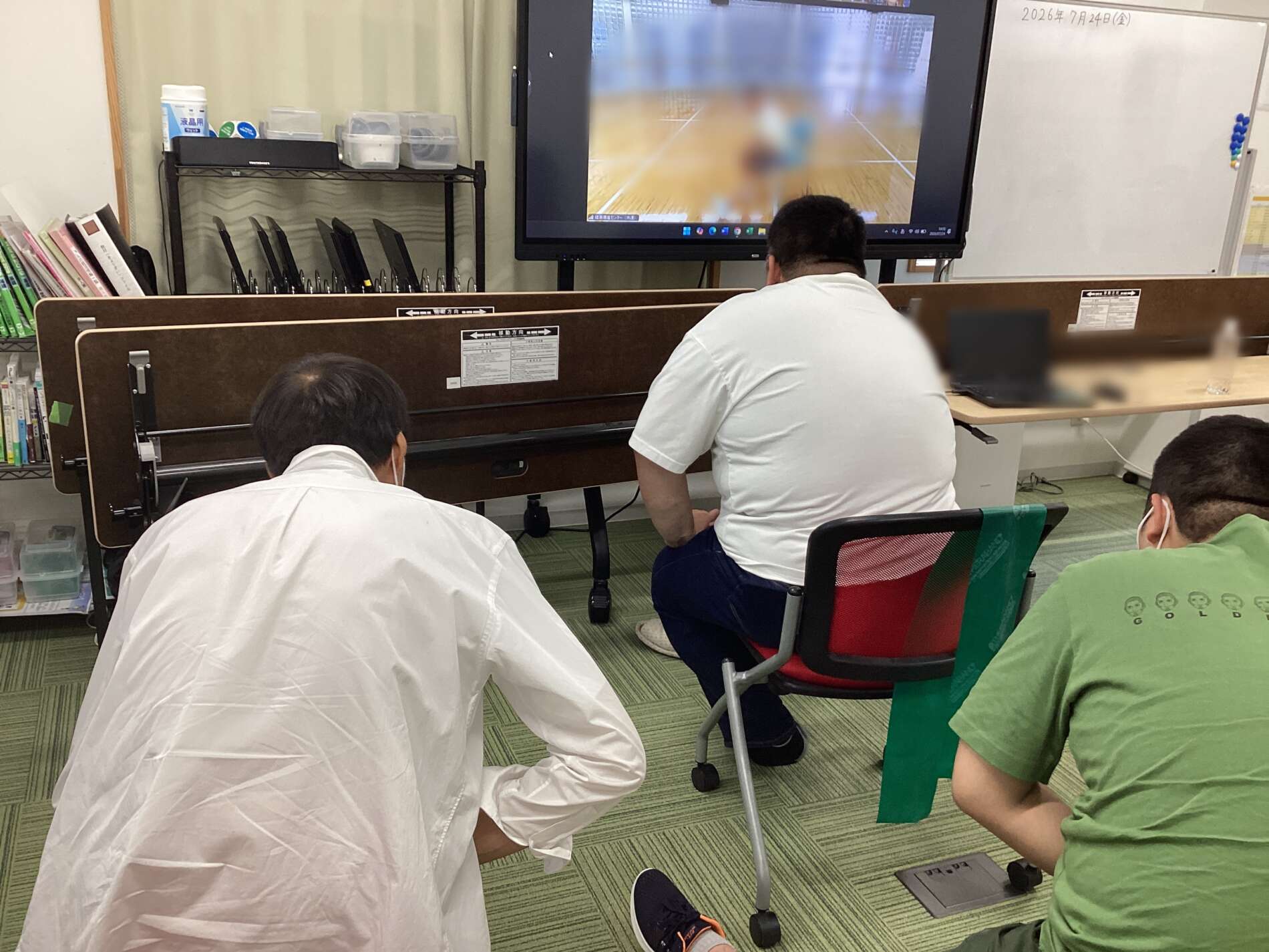 Students in a classroom seated behind brown partitions, watching a large screen showing a blurred sports court.