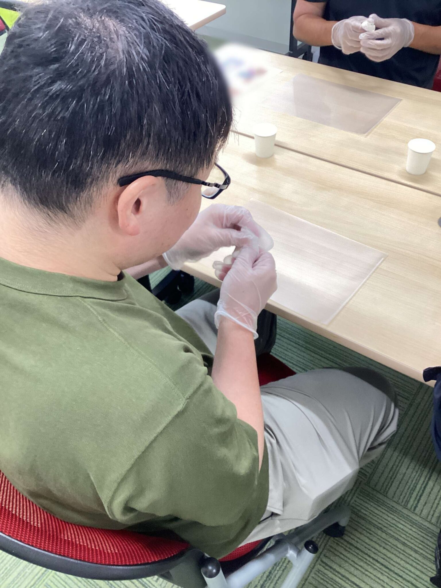 Person in a green shirt sits at a table wearing gloves and handling a small item; another gloved person stands in the background near a protective sheet on the table