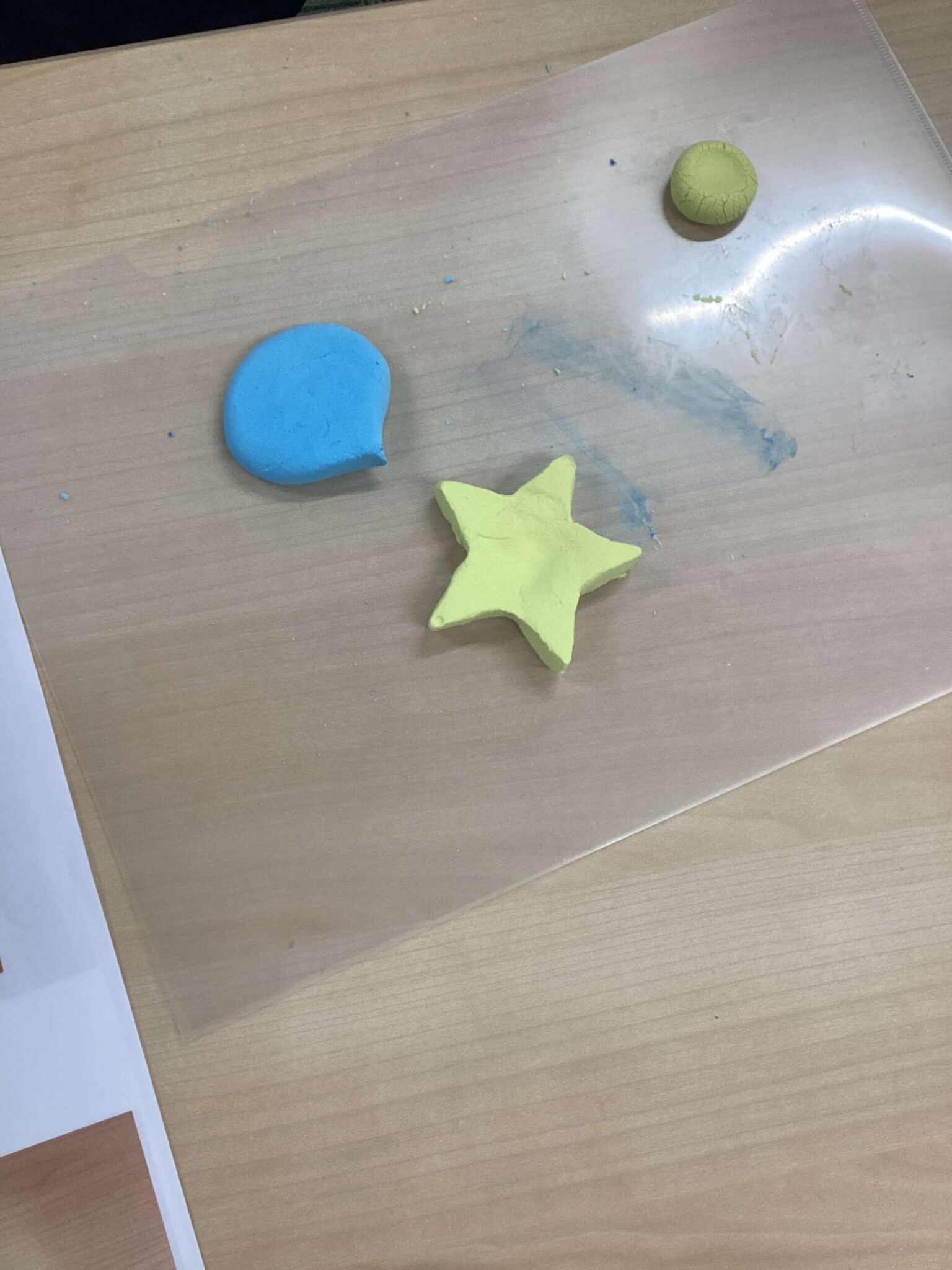 Blue clay blob and a yellow star-shaped piece, with a small green clay ball on a wooden table, plus paint smudges in the background — an arts-and-crafts scene.