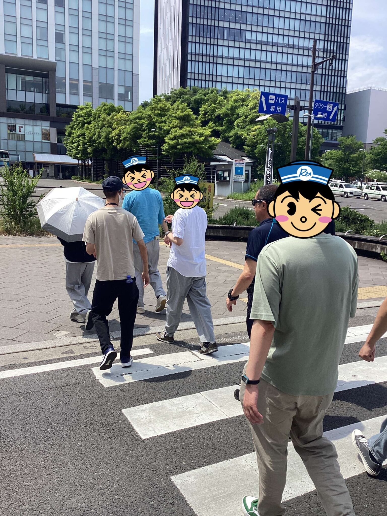 People crossing a city street at a crosswalk, with modern office buildings and green trees in the background; one person holds a white umbrella.
