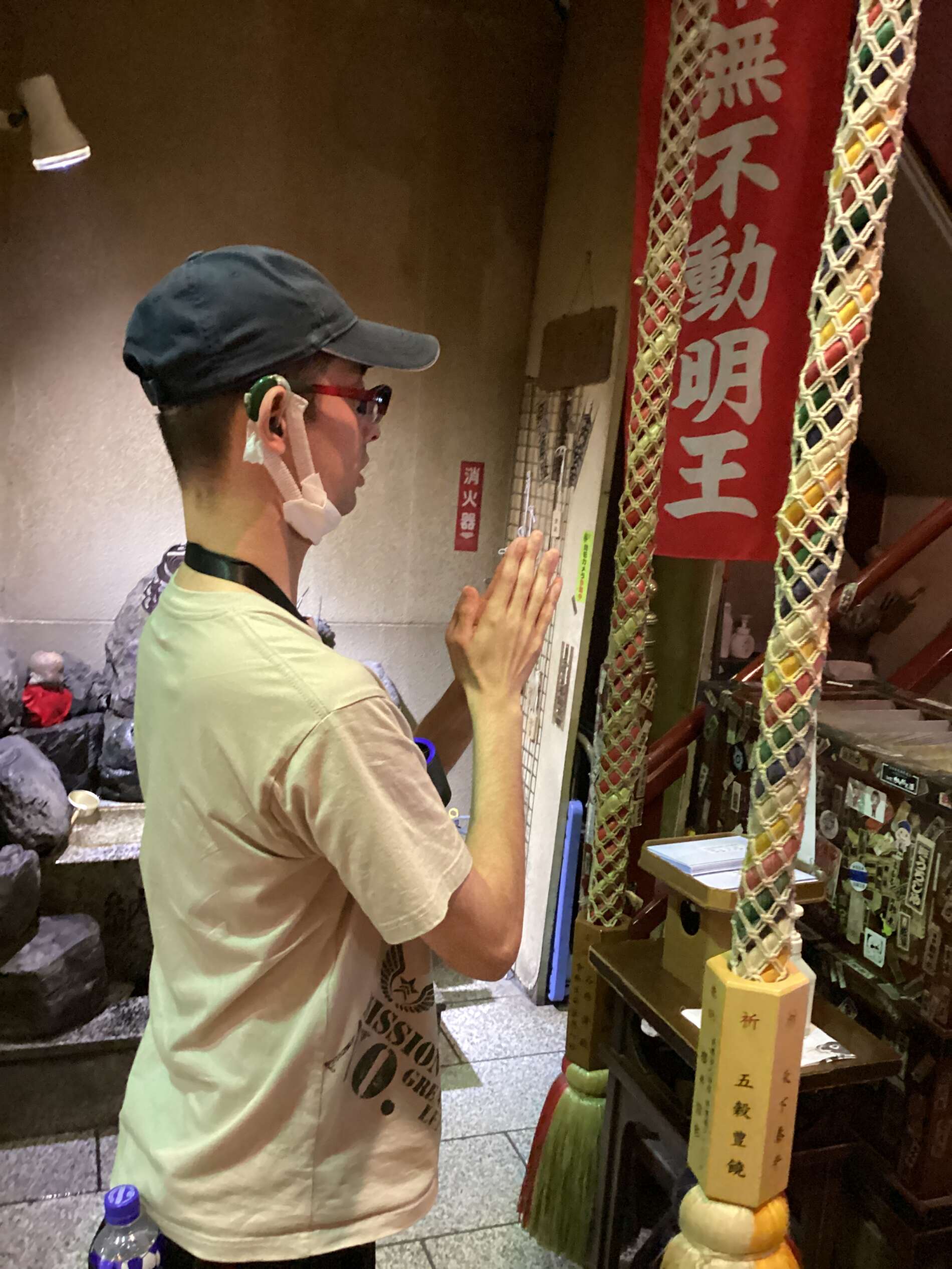 Man in a beige shirt and cap prays with hands together at a Japanese shrine, red banner and braided rope visible behind him.