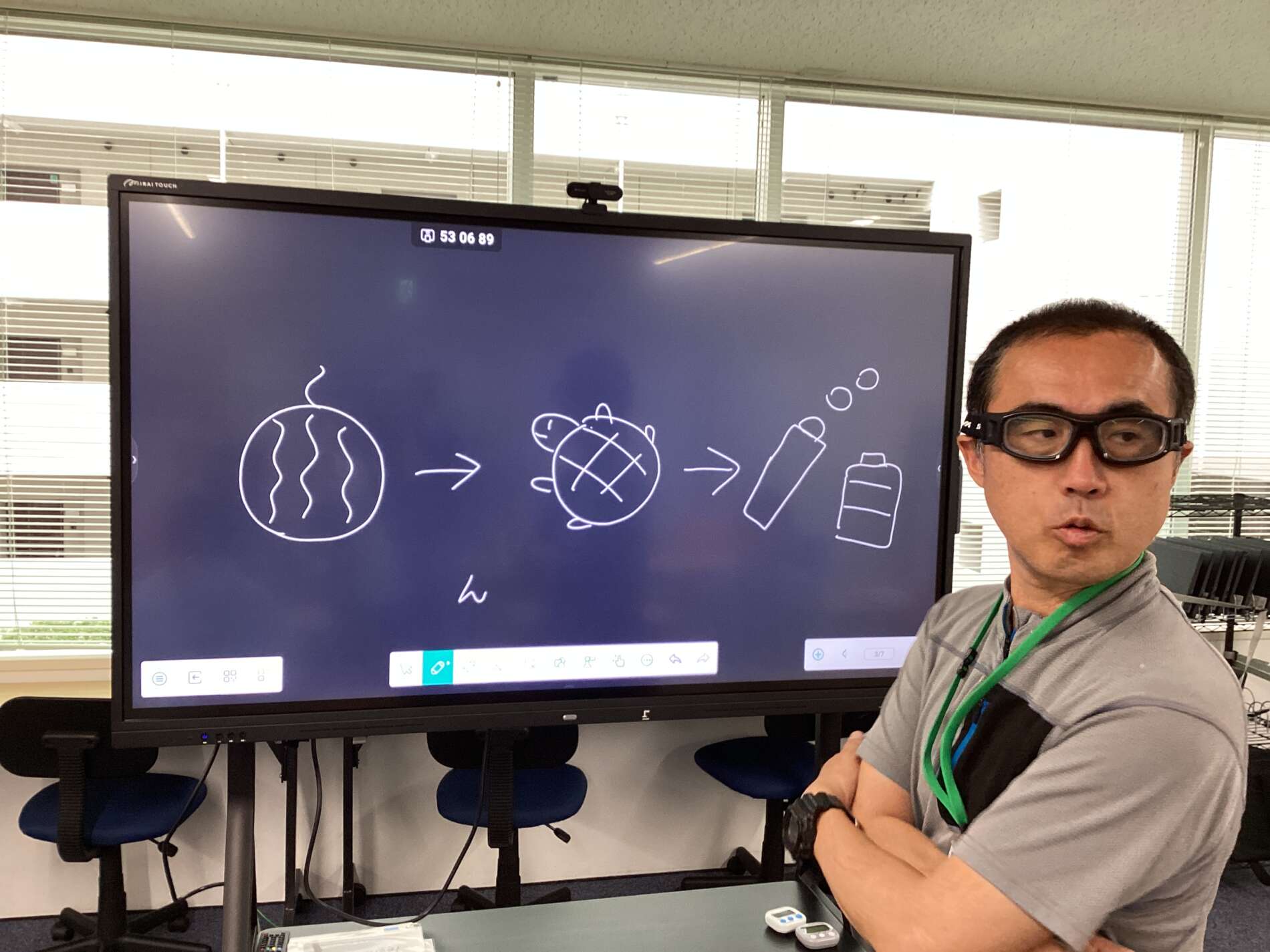 Instructor in safety goggles beside a large interactive screen showing hand-drawn science icons: a round melon-like circle, a globe, a bottle, and a container.