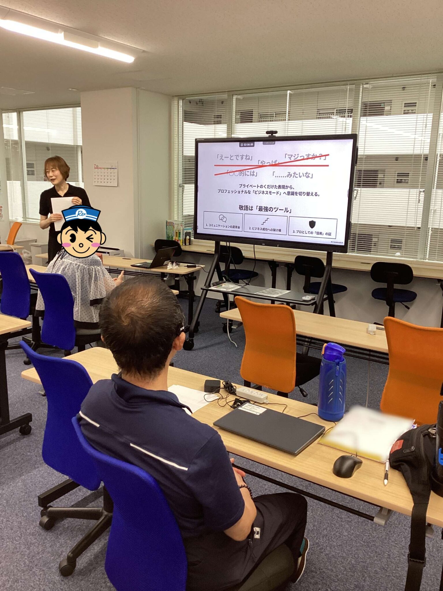 Instructor standing at the front of a classroom presenting to seated attendees; a large screen shows a Japanese slide deck with red underlines and diagrams, while students sit at desks with notebooks and laptops.
