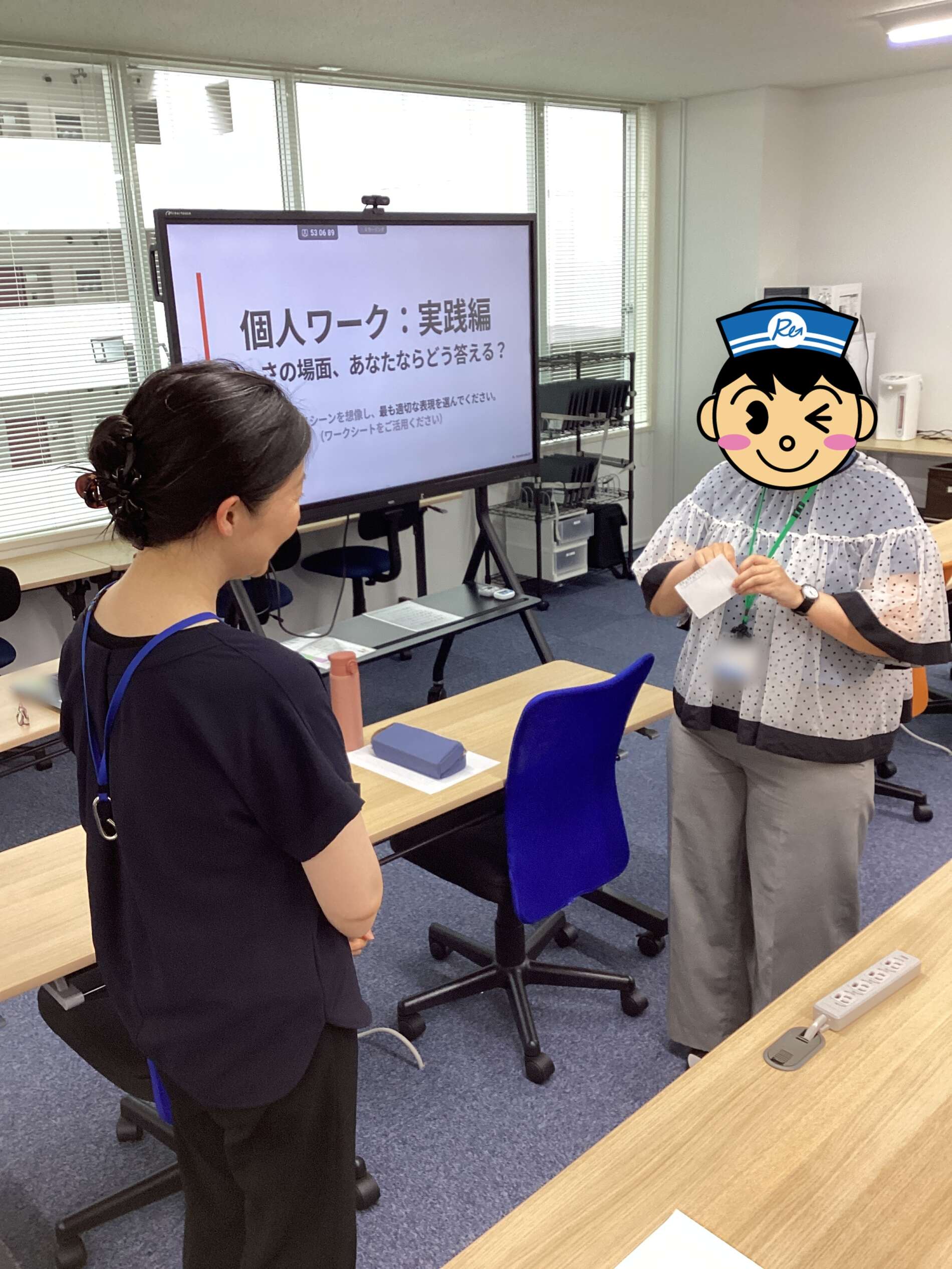 Office classroom scene with a large presentation screen showing Japanese text, two people near desks discussing.