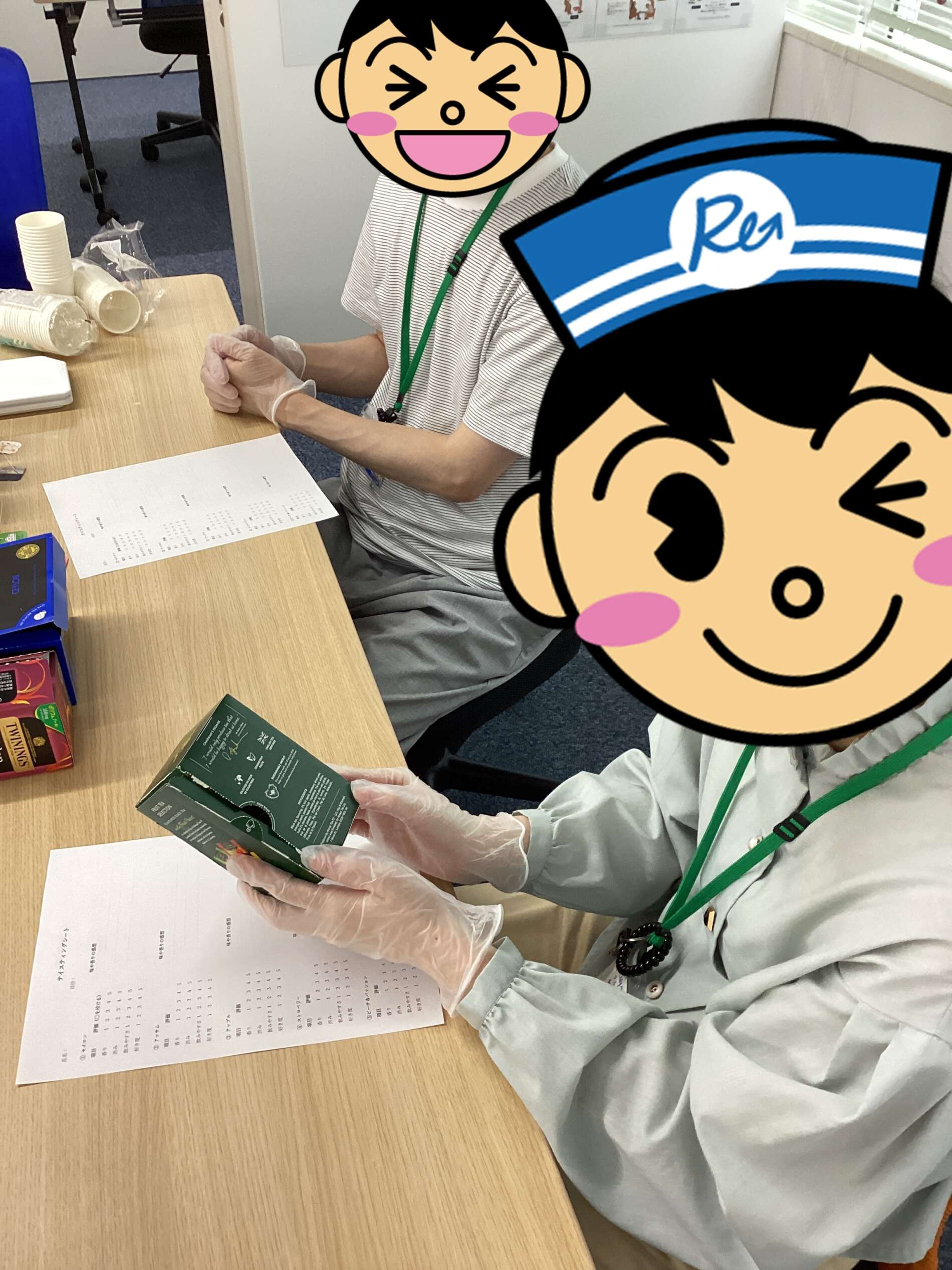 Two people wearing gloves and green lanyards sit at a wooden table, handling a green box and a printed sheet, with disposable cups and snack boxes nearby—office or lab setting.