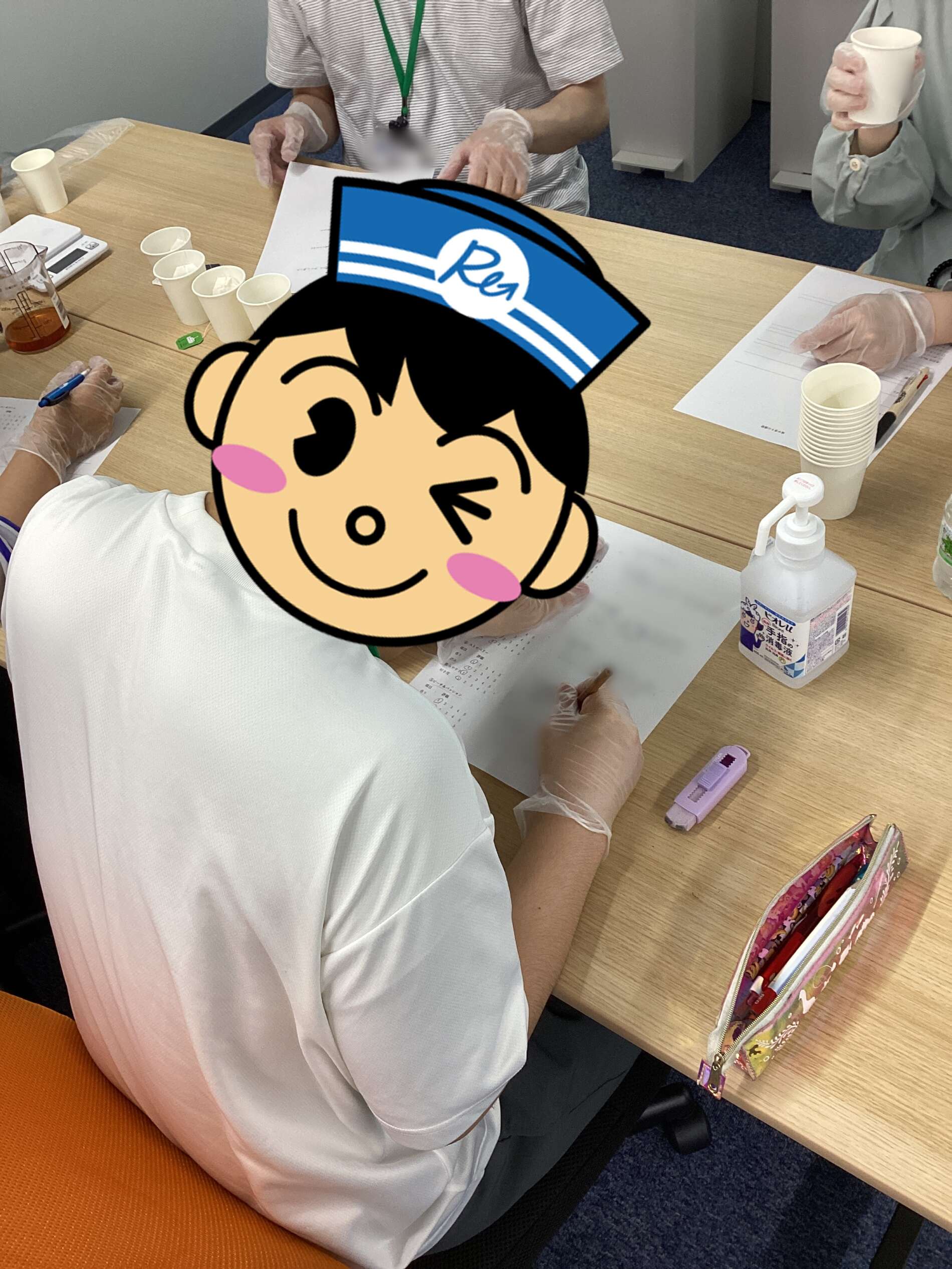 Person wearing gloves signs a form at a wooden table with cups and a sanitizer bottle nearby, a cartoon face sticker covers part of the view for privacy.