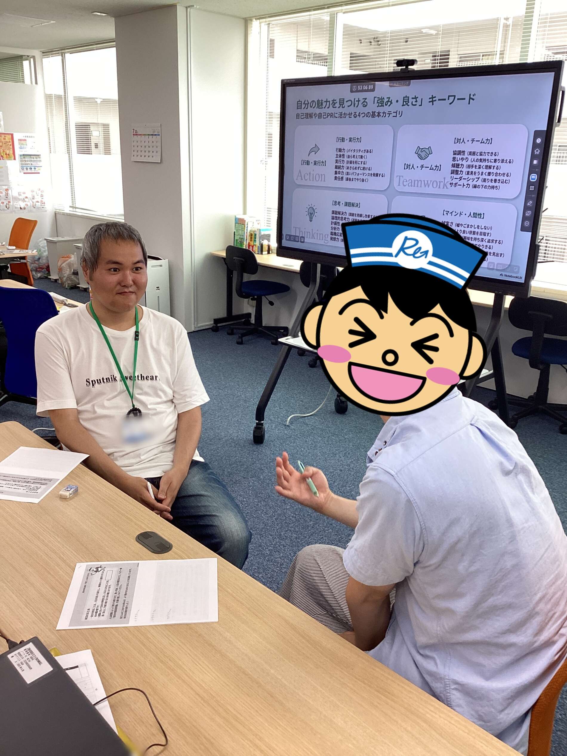Two people sit across from each other at a office table, discussing with papers on the table and a large presentation screen in the background.