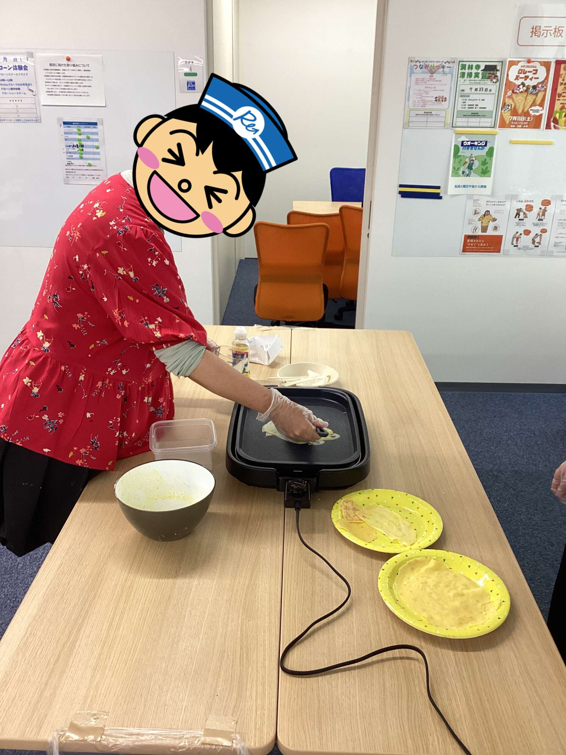 Person in a red floral blouse cooking pancakes on a black electric skillet at a table, with yellow plates of crepes nearby.