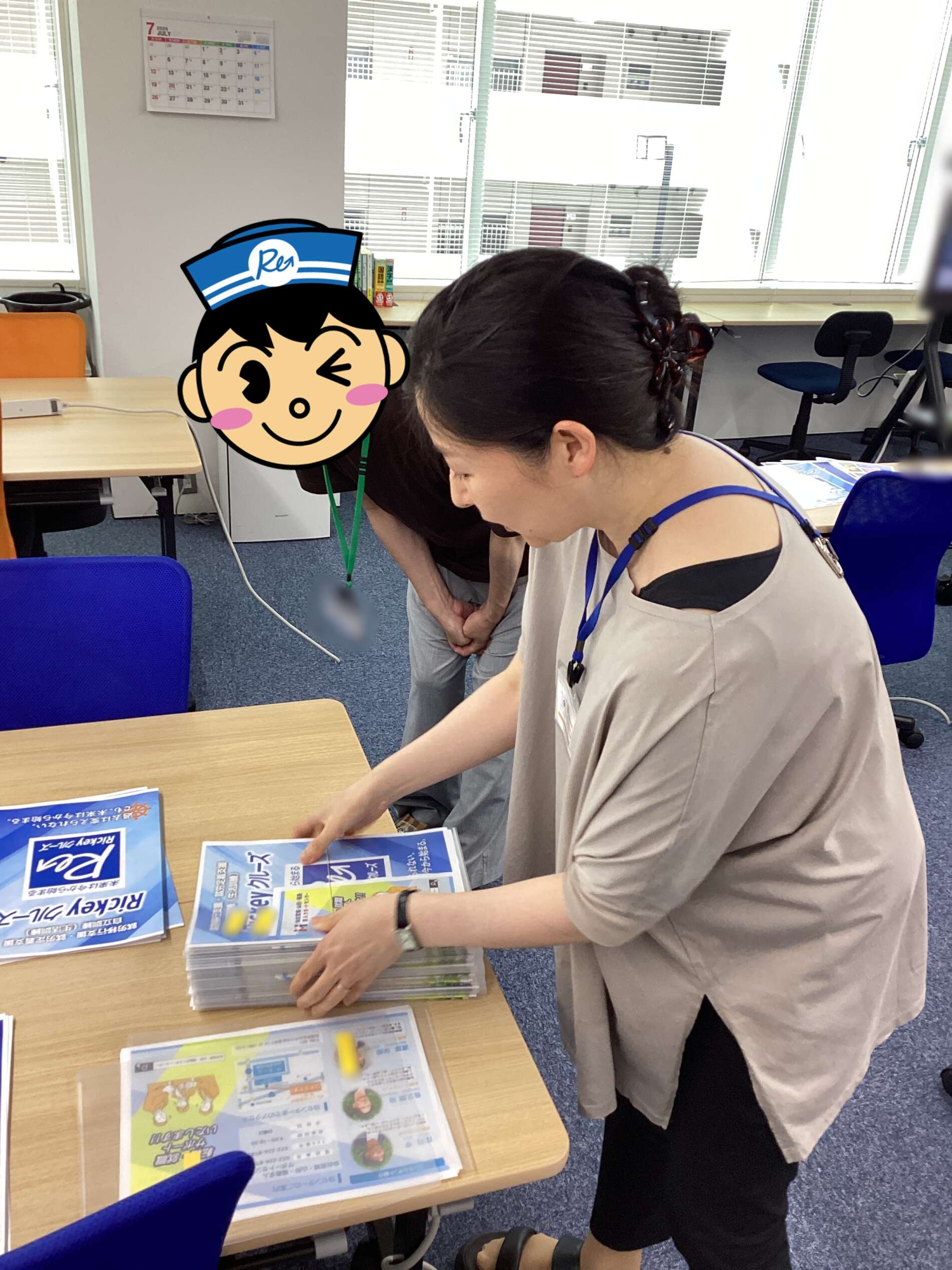 Person in a beige top sorts blue-and-yellow brochures on a desk in a classroom, with a cartoon cutout obscuring another person’s face; blue chairs and a calendar on the wall in view.