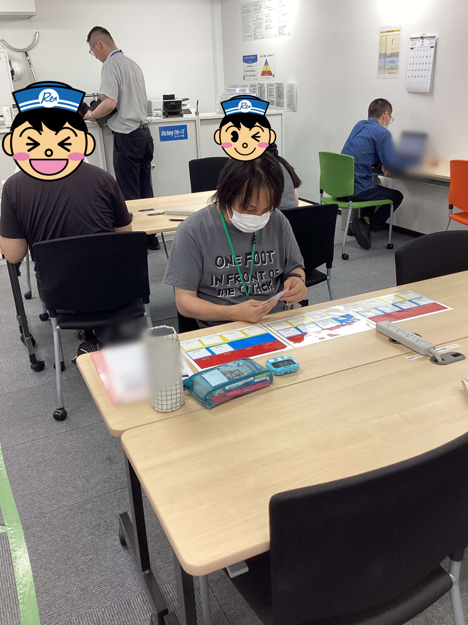 People seated at tables in a classroom or training room; one person at the front studies papers while others work, masks worn, colorful cards spread on the desk.