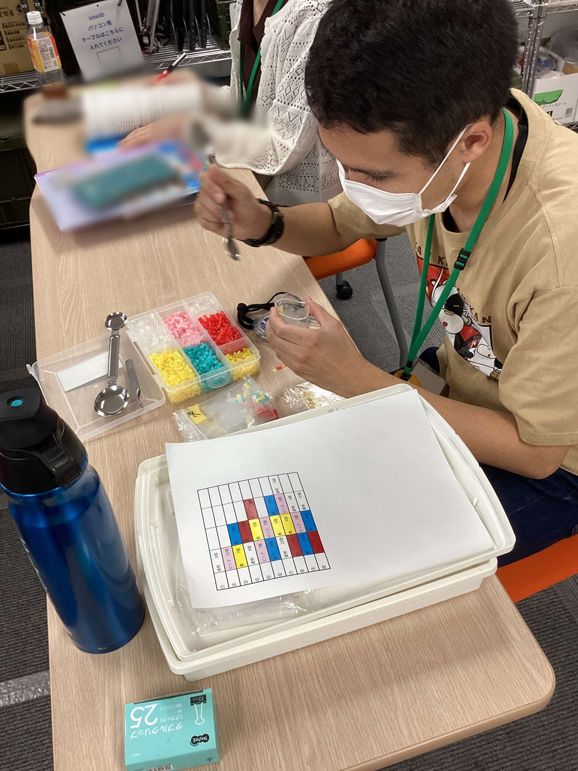 Person wearing a mask and green lanyard sits at a desk sorting small colorful beads into a plastic bead organizer; a color chart lies on the tray, with a blue water bottle nearby in a workshop or classroom setting.