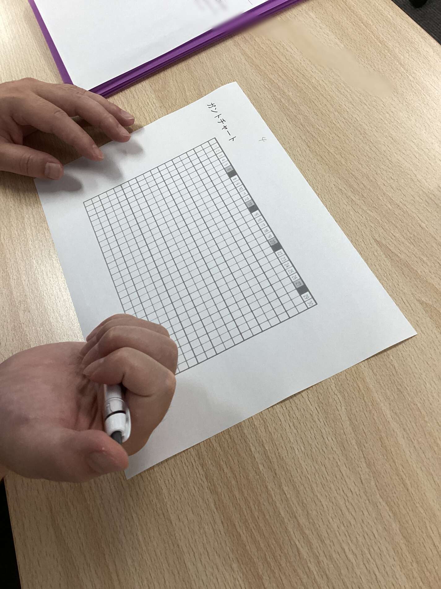Person's hands hold a pen over a sheet of graph paper on a wooden desk, ready to write; a purple-bound notebook sits nearby.