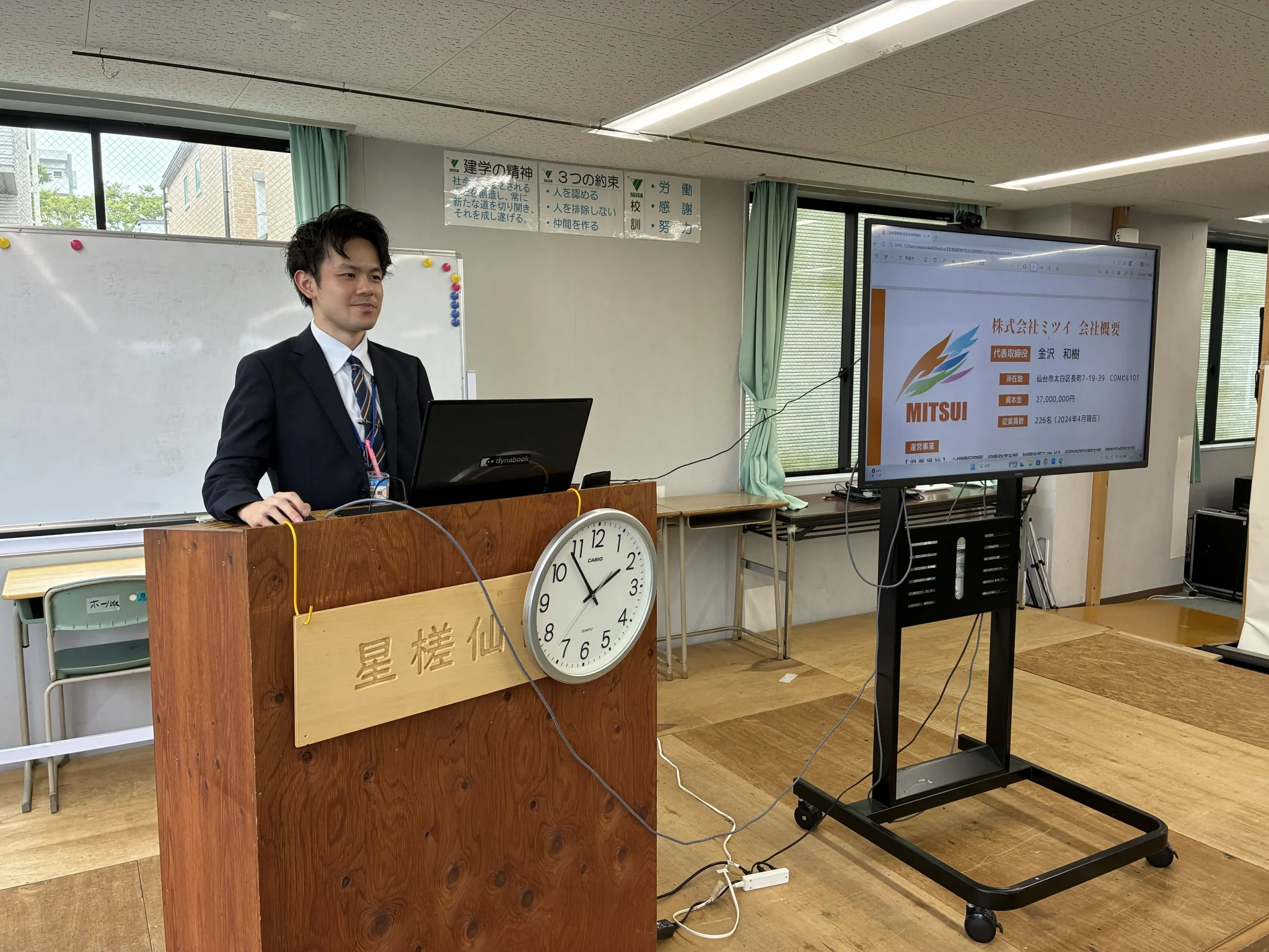 Man in a dark suit giving a presentation at a wooden podium in a classroom, with a large wall clock and a digital screen showing a Mitsui slide.