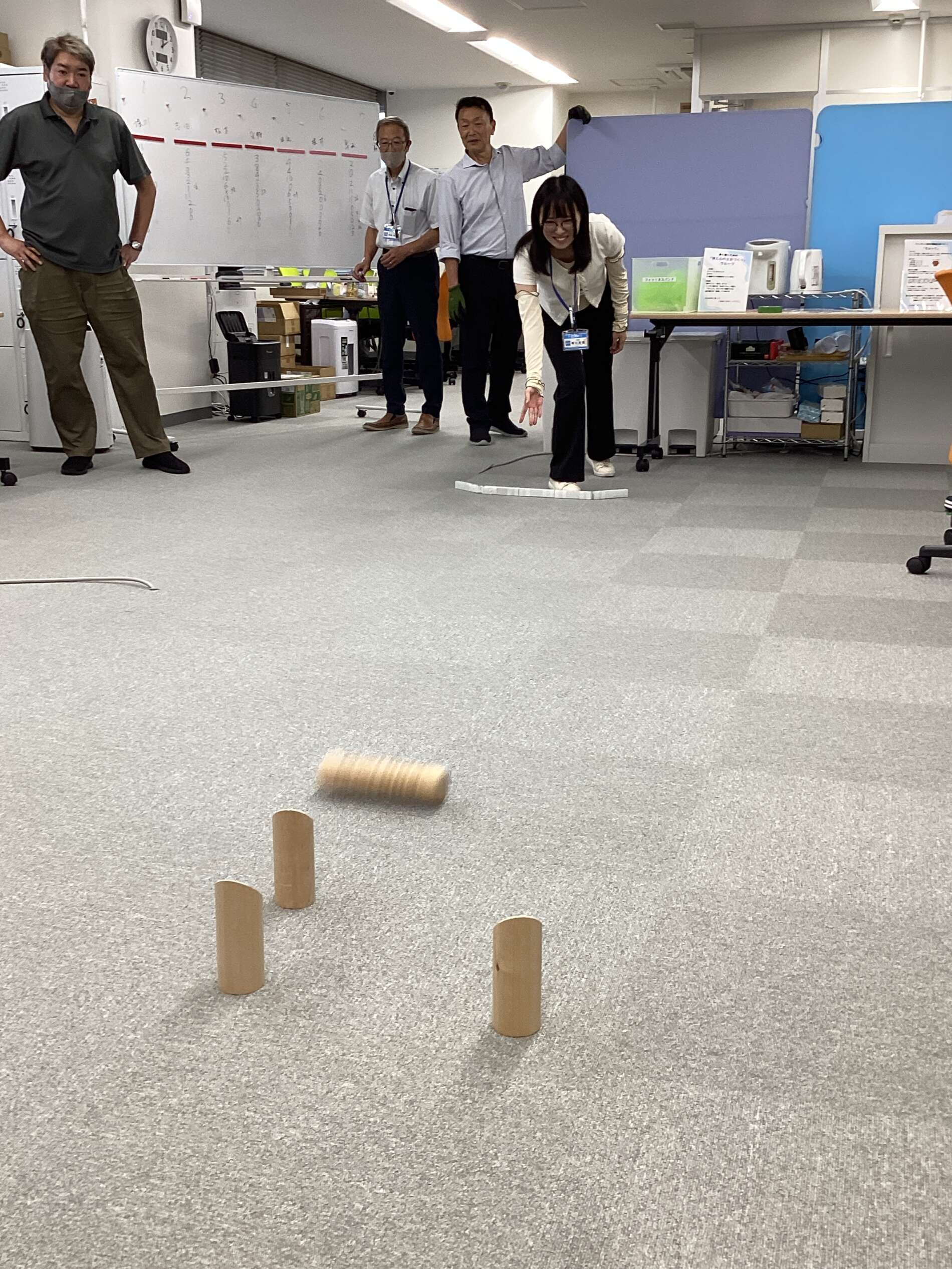 Woman in white top balances on a curved row of white blocks on the office carpet while colleagues look on near a whiteboard and tables.