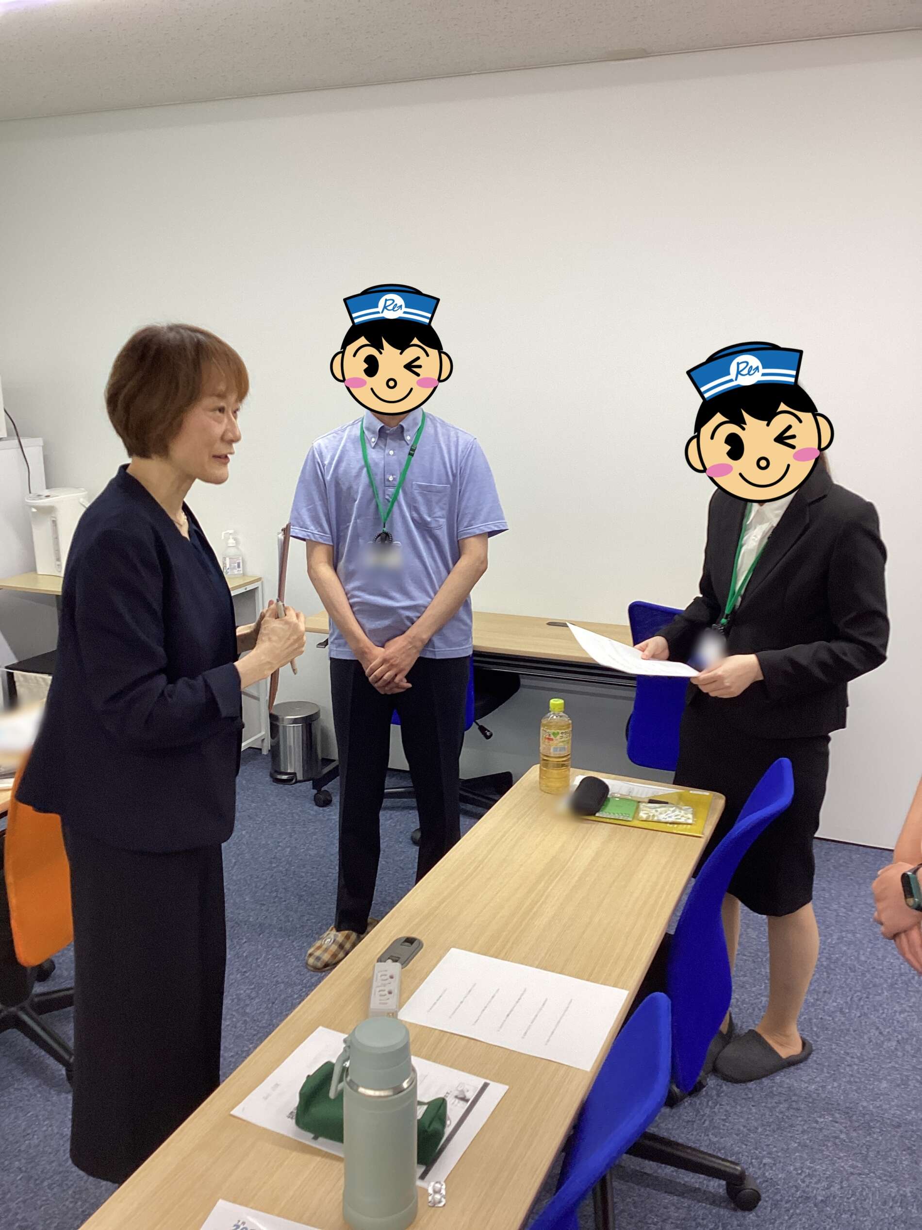 Three colleagues stand around a long office table, one woman speaking while two men listen, with papers and a bottle on the table; faces covered by cartoon icons for privacy.