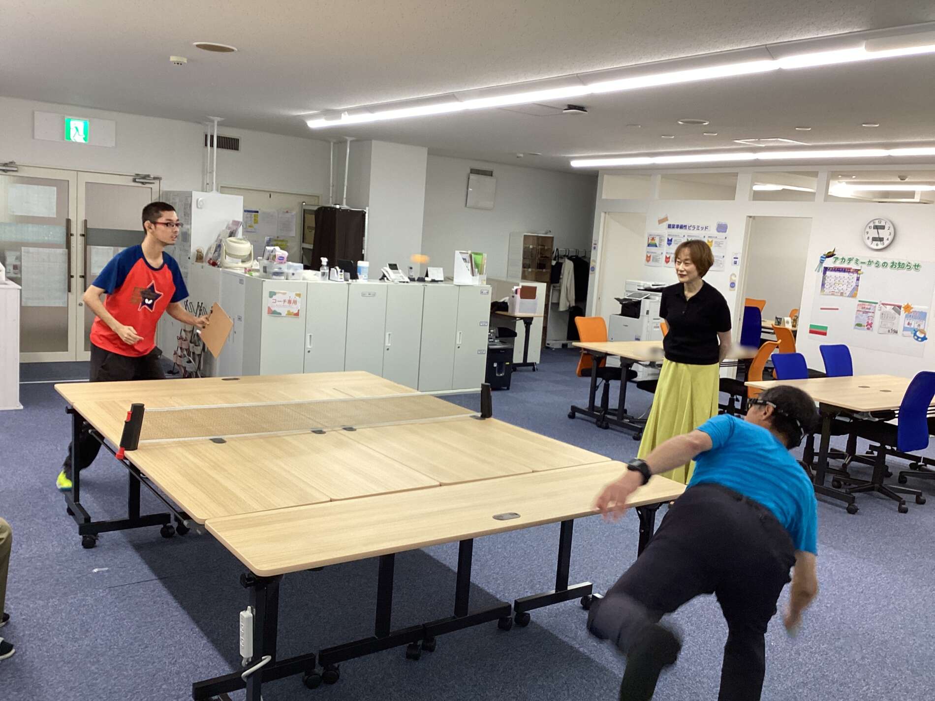 People playing table tennis in a bright office-like room with several tables and chairs around them.