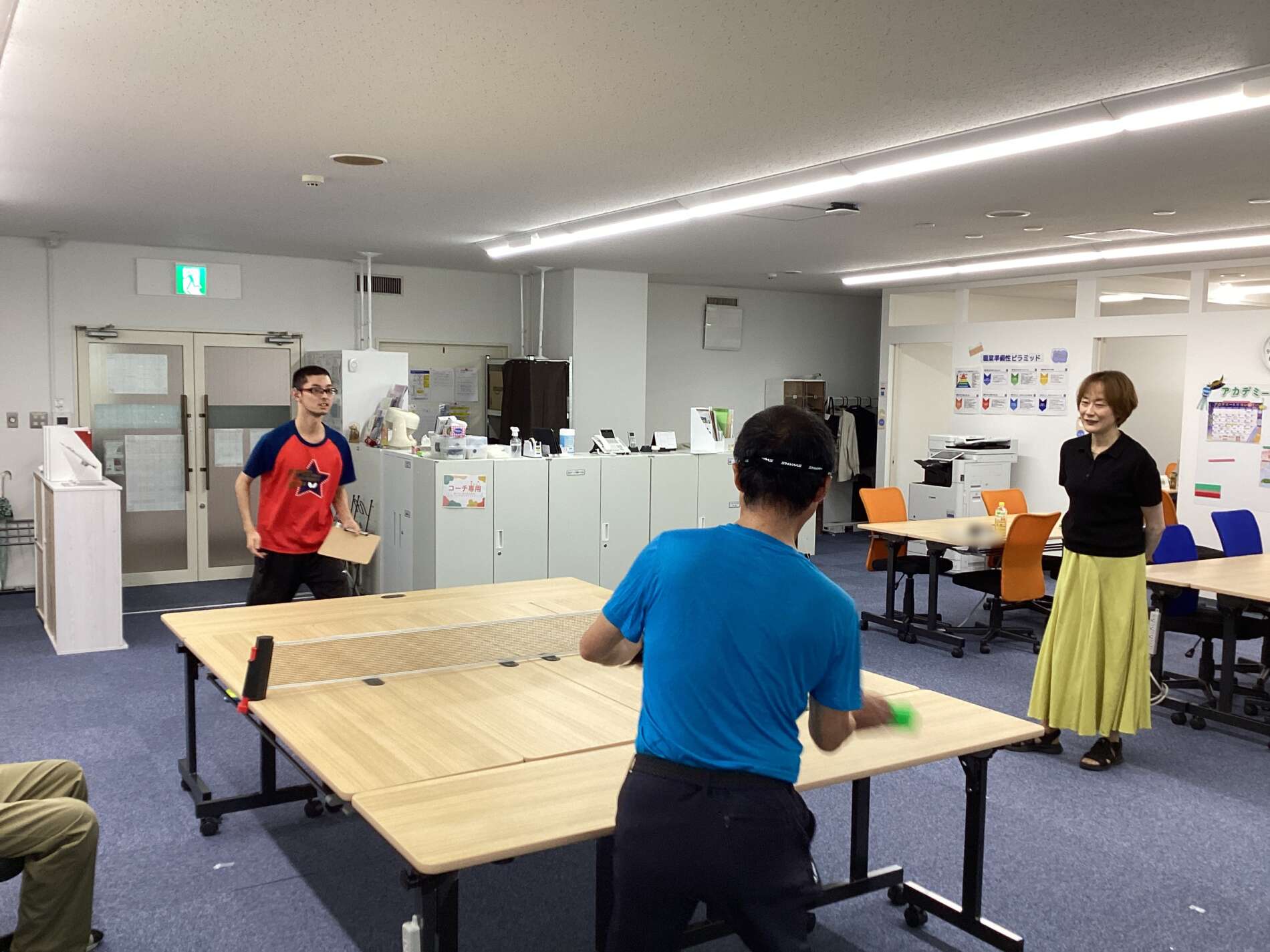 People playing table tennis in a bright room; a man in a blue shirt serves at the near table while others stand around.