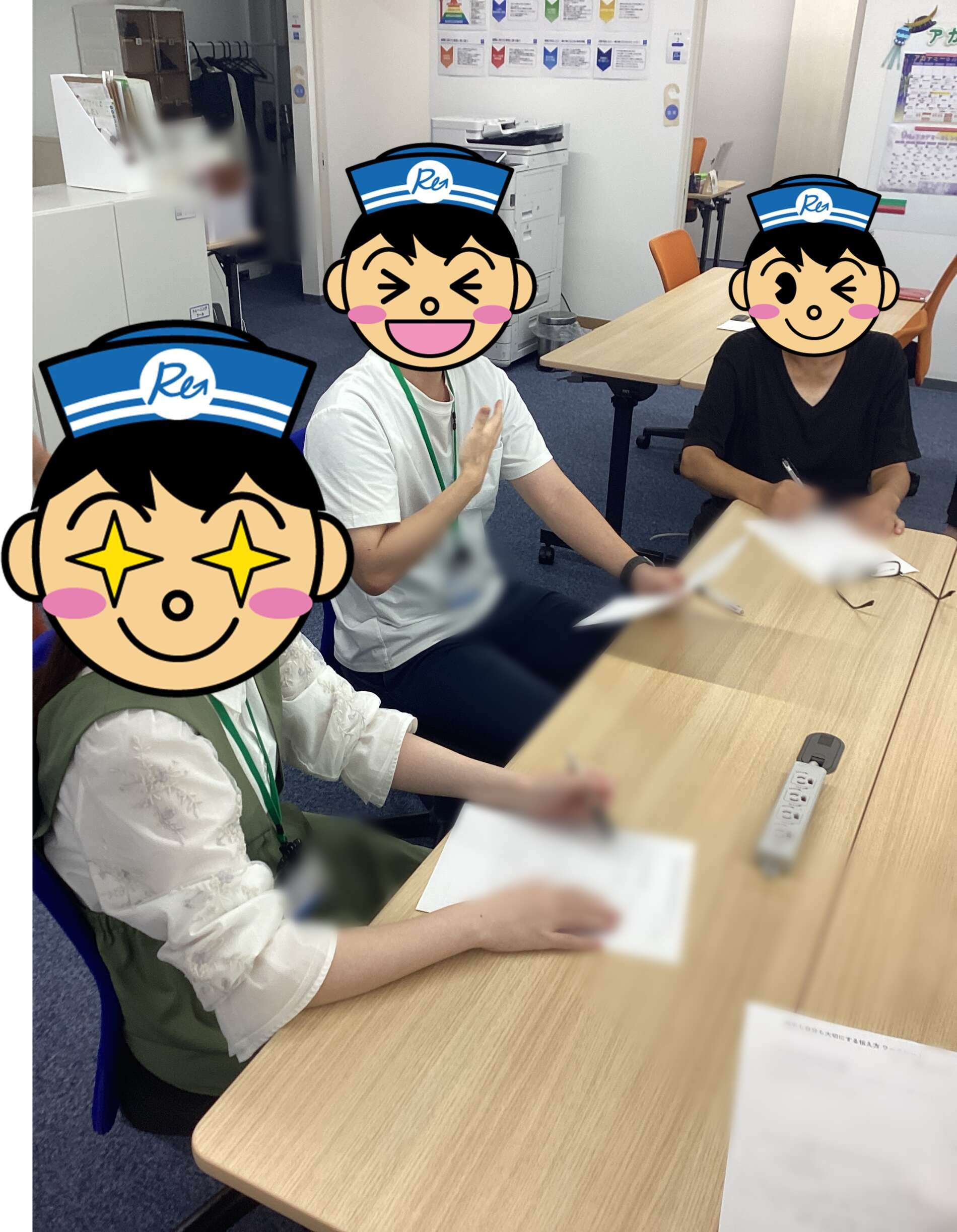 Three people sit at a rectangular table in a classroom or office, wearing cartoon nurse masks; one person writes while others look on. In the background are posters and office equipment.