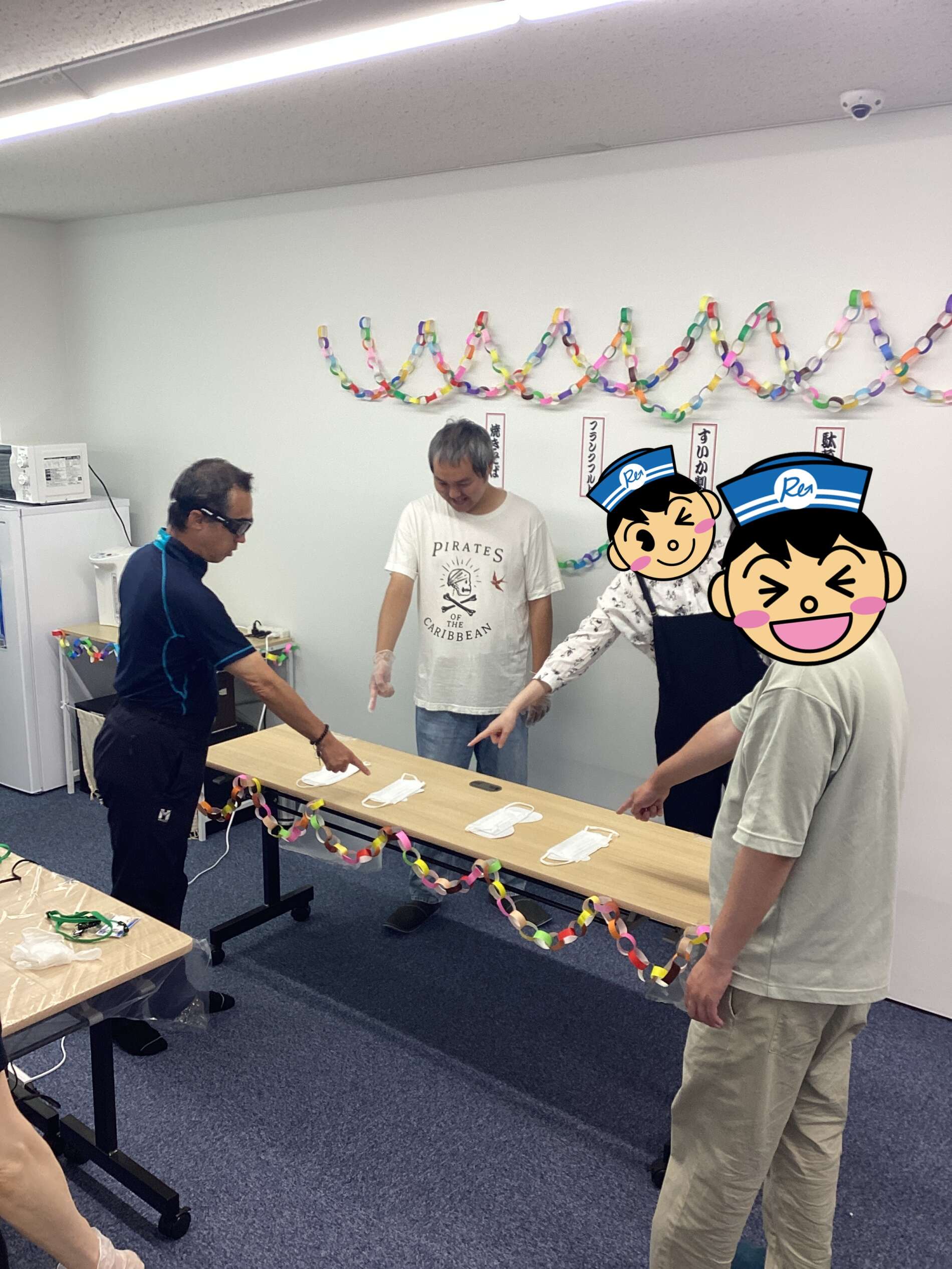 People in a room around a long table assembling face masks, with colorful rainbow paper-chain garlands on the wall behind them (gloves on).