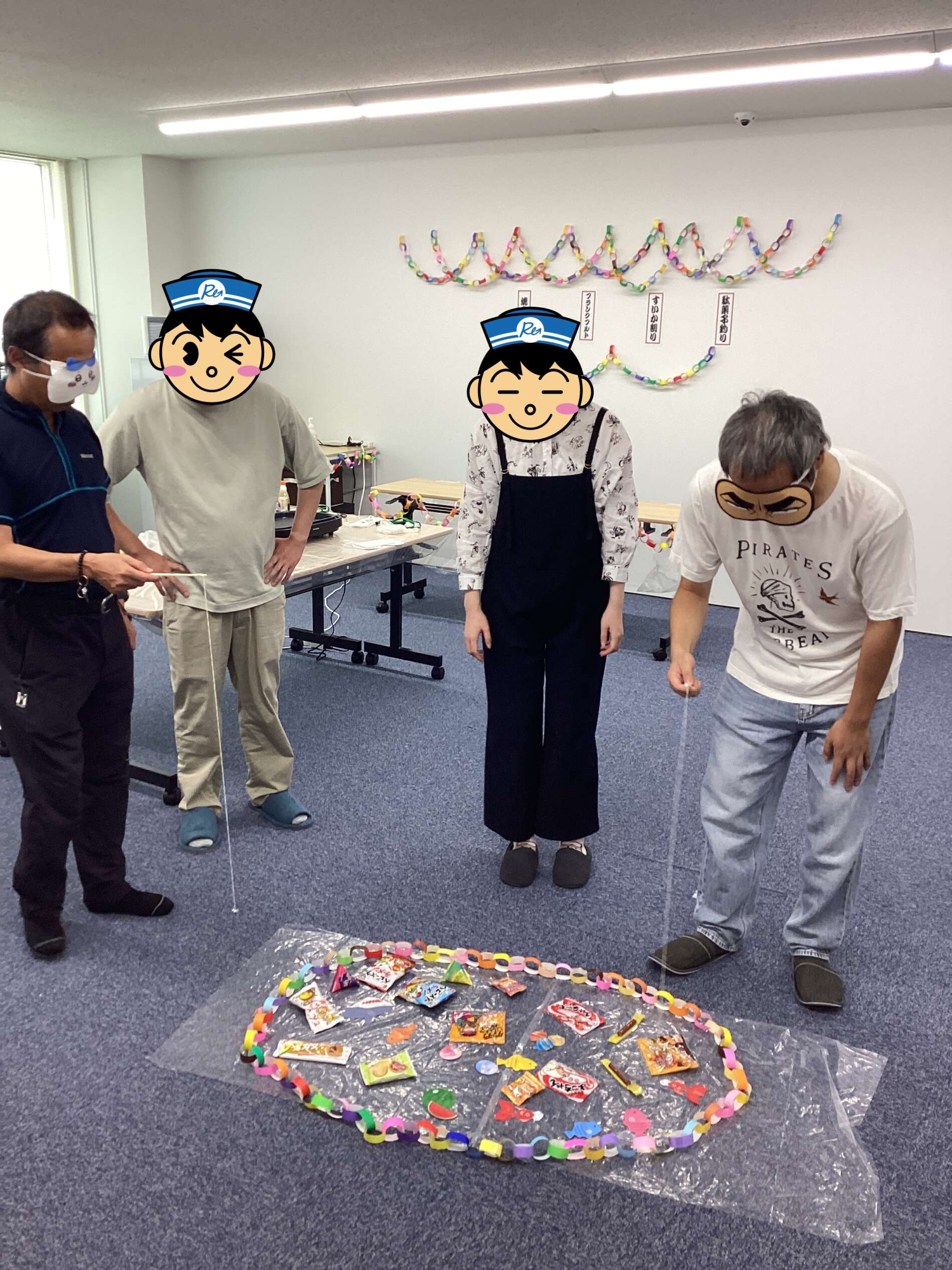 Four people wearing face masks stand around a colorful paper-chain display and scattered snacks on the floor, as if taking a break in a room decorated with a chain garland in the background.
