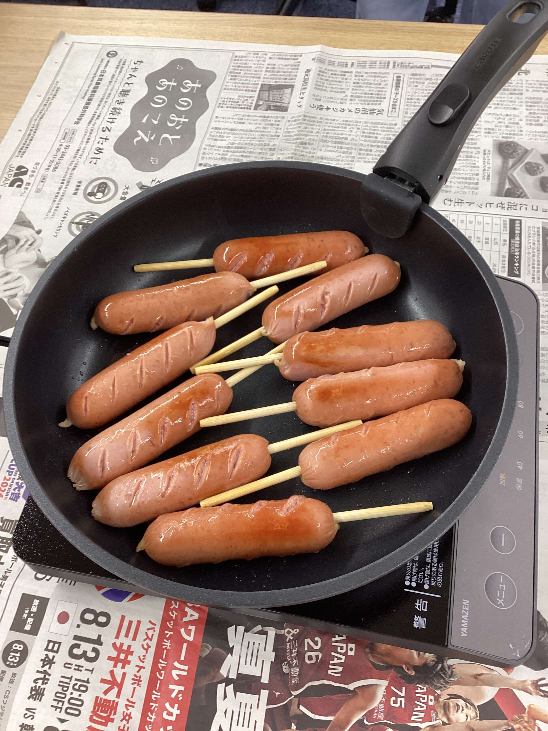 Pan with several grilled sausages on wooden skewers in a black skillet, resting on a newspaper-covered surface.