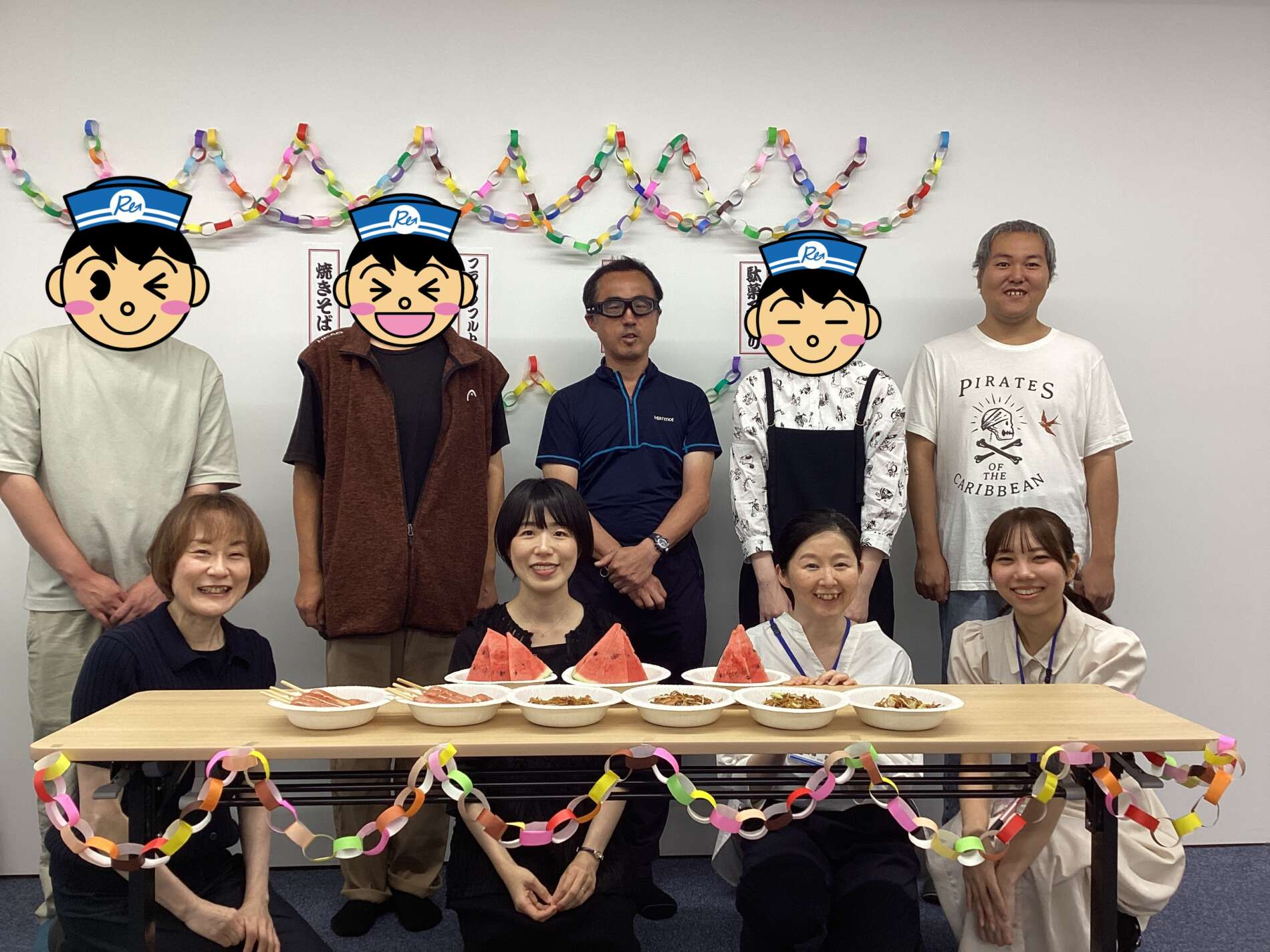 Group of people posing at a celebration around a table with plates of food and watermelon slices, with colorful paper garlands in the background.